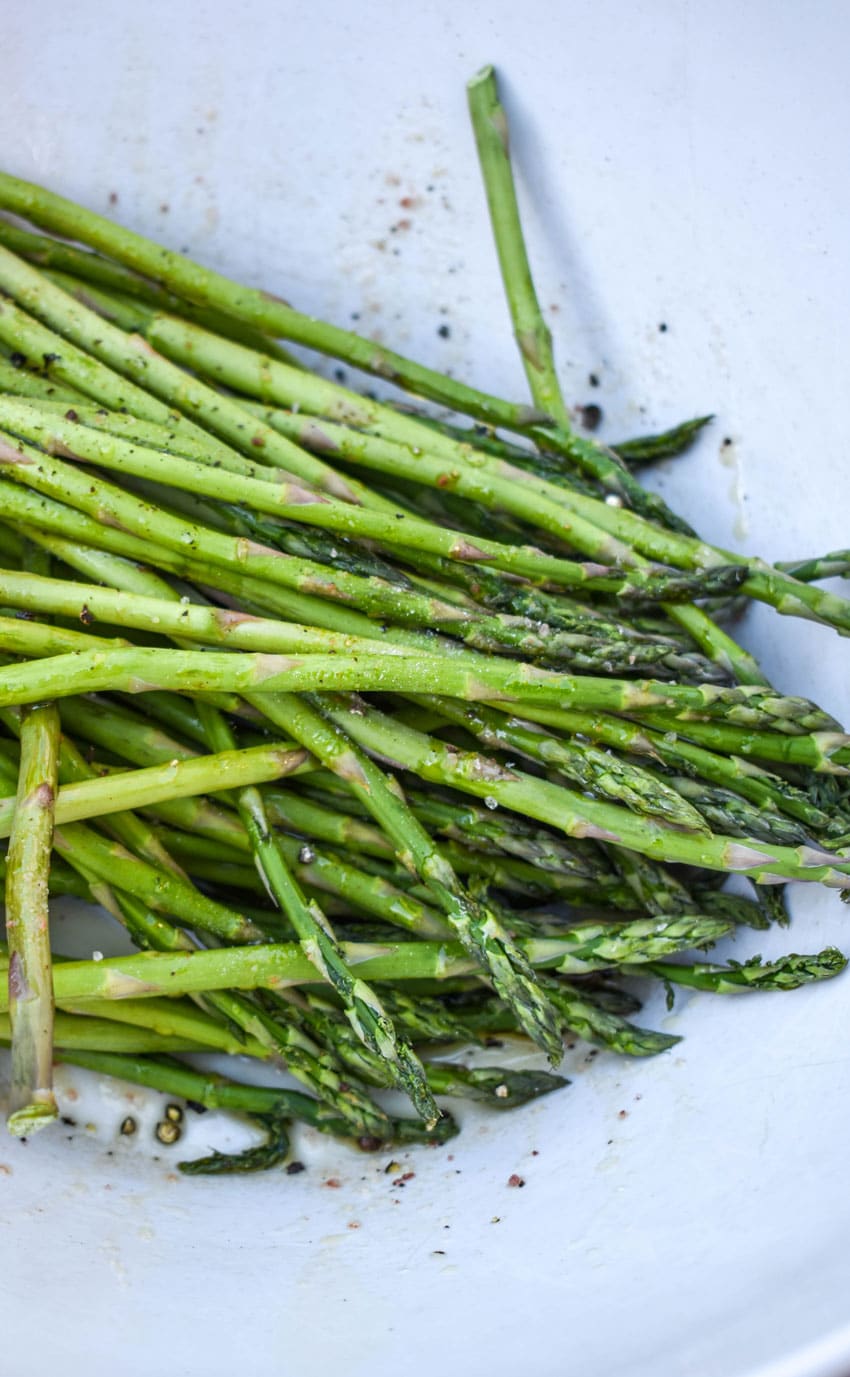 stalks of asparagus coated in olive oil and seasoned with salt in pepper in a white mixing bowl