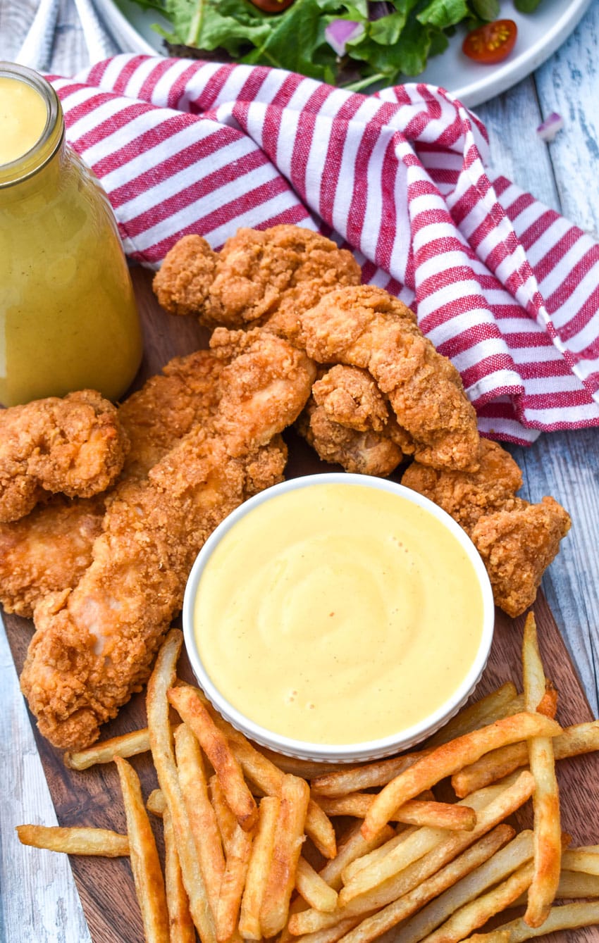 homemade honey mustard sauce in a small white dipping bowl surrounded by chicken tenders and french fries