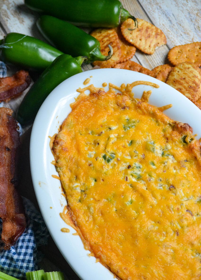 jalapeno popper di in a white serving dish surrounded by strips of crisp bacon, jalapeno peppers, and pretzel crackers
