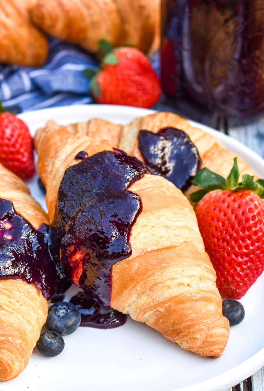 a close up of crockpot blueberry sauce spread over two croissants on a white plate with fresh fruit on the side