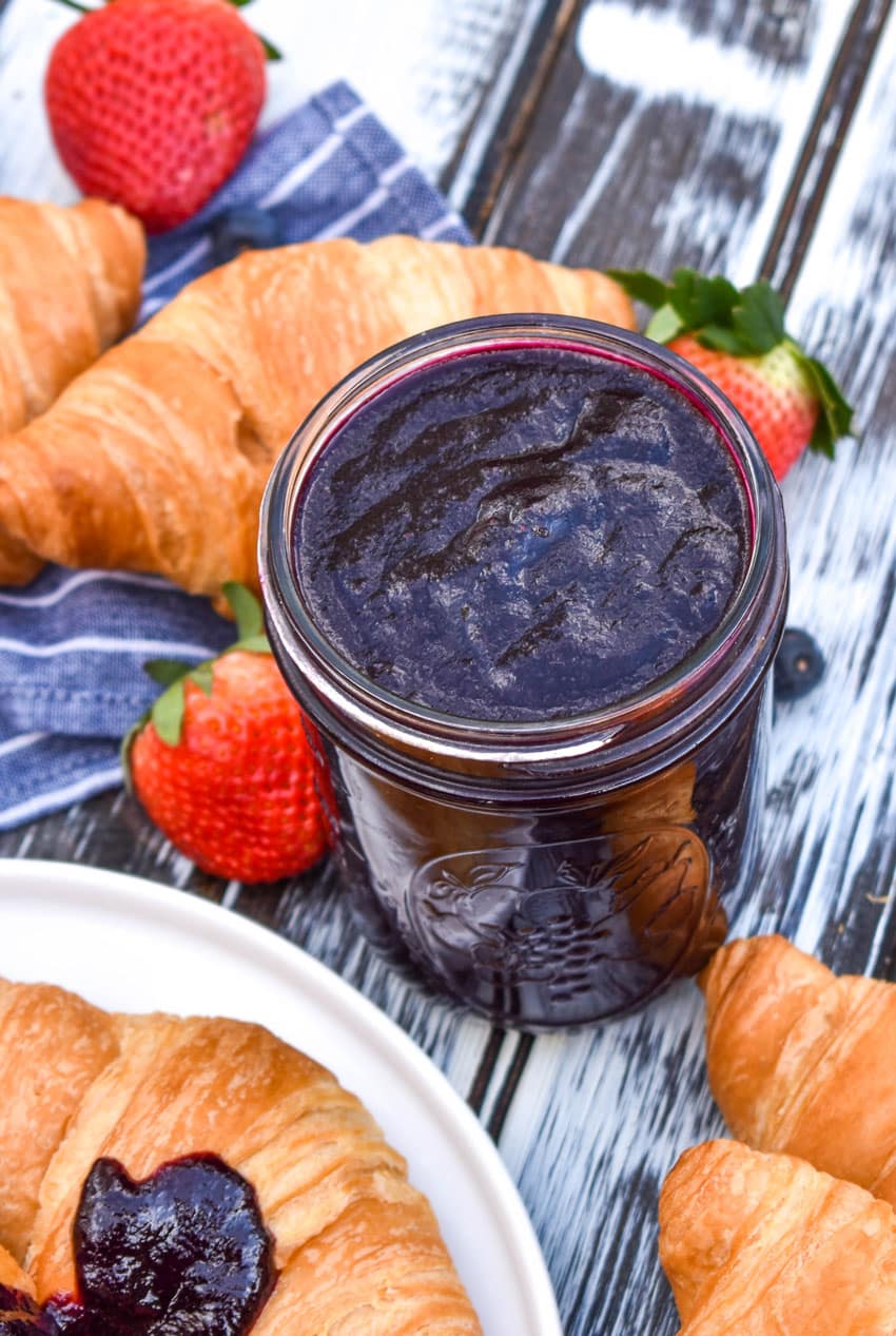 slow cooker blueberry butter in a glass jar surrounded by fresh fruit and croissants