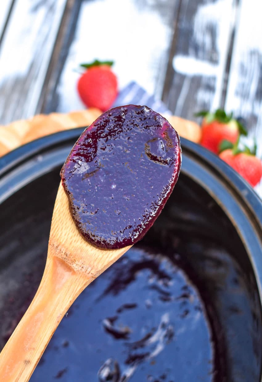 a wooden spoon holding a scoop of slow cooker blueberry butter over a black crock