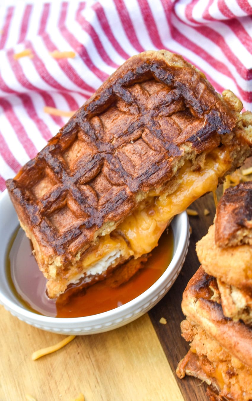 half of a chicken and waffle grilled cheese being dipped in a small white bowl filled with maple syrup
