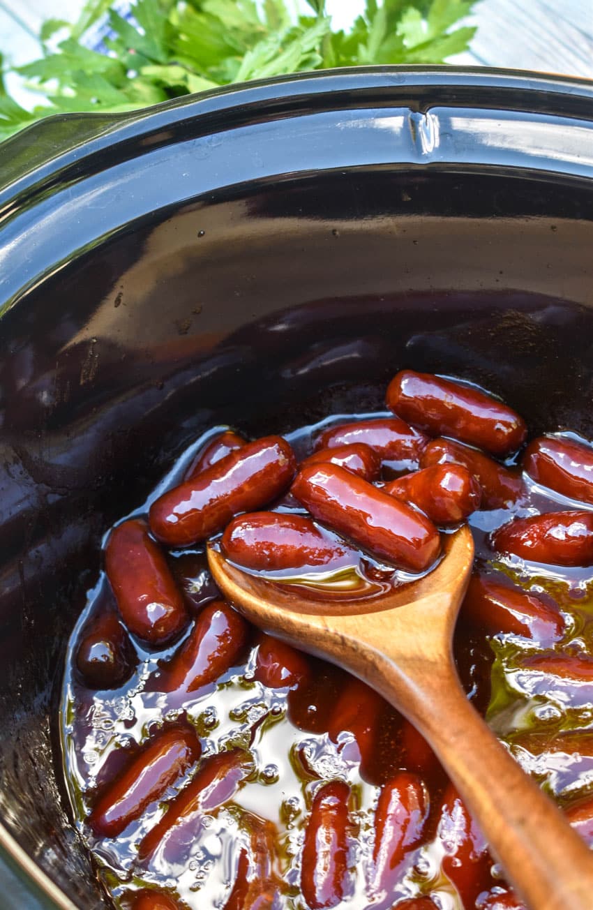 a wooden spoon scooping slow cooker little smokies out of the black bowl of a crockpot