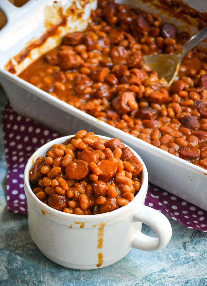 hotdogs and beans casserole in a white mug next to a white baking dish
