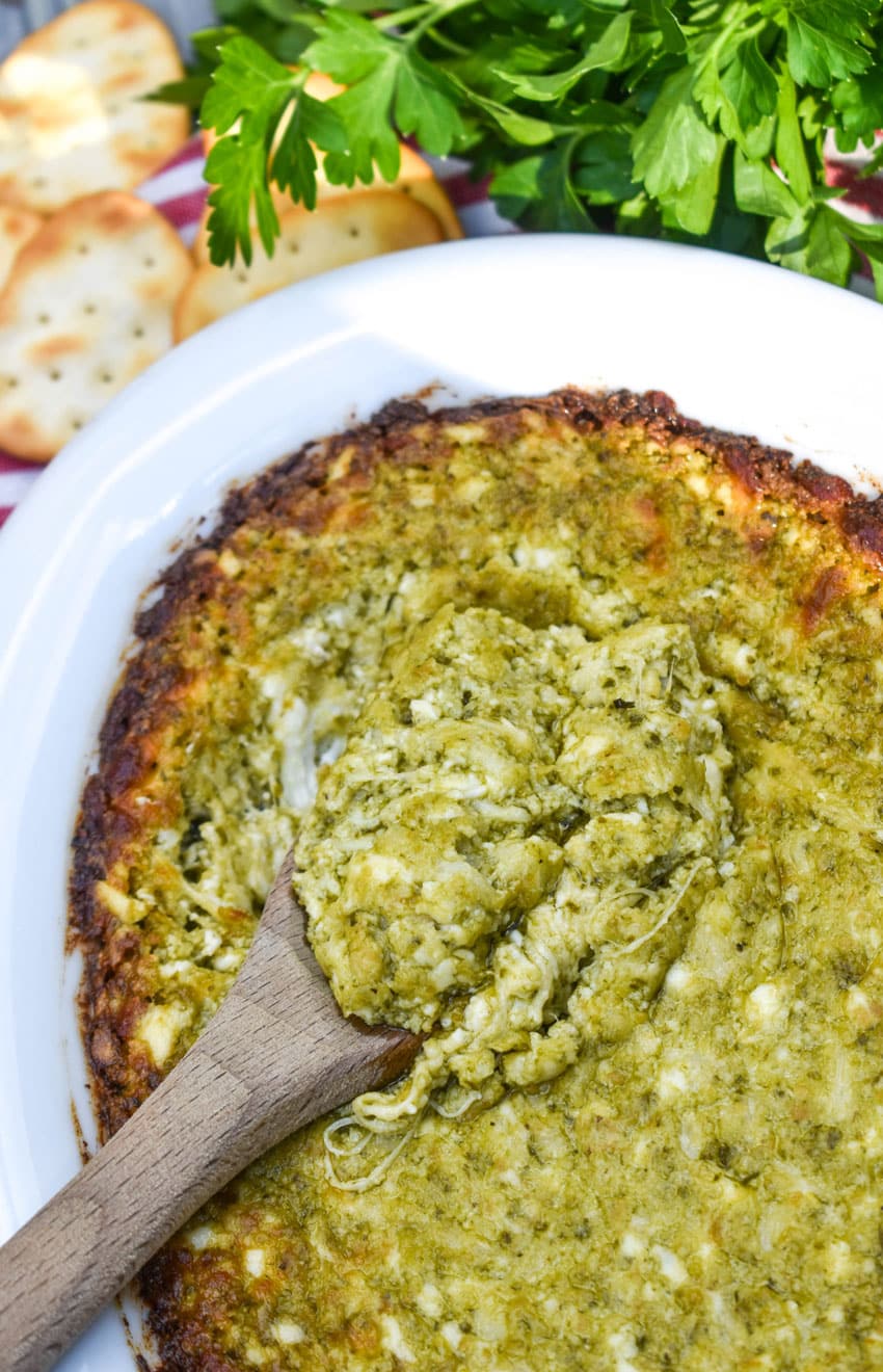 a wooden spoon scooping hot pesto dip out of a white serving dish