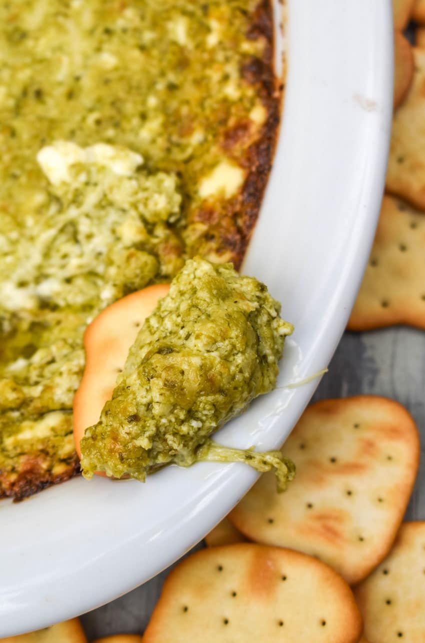 a cracker topped with hot pesto dip resting on the edge of a white serving dish