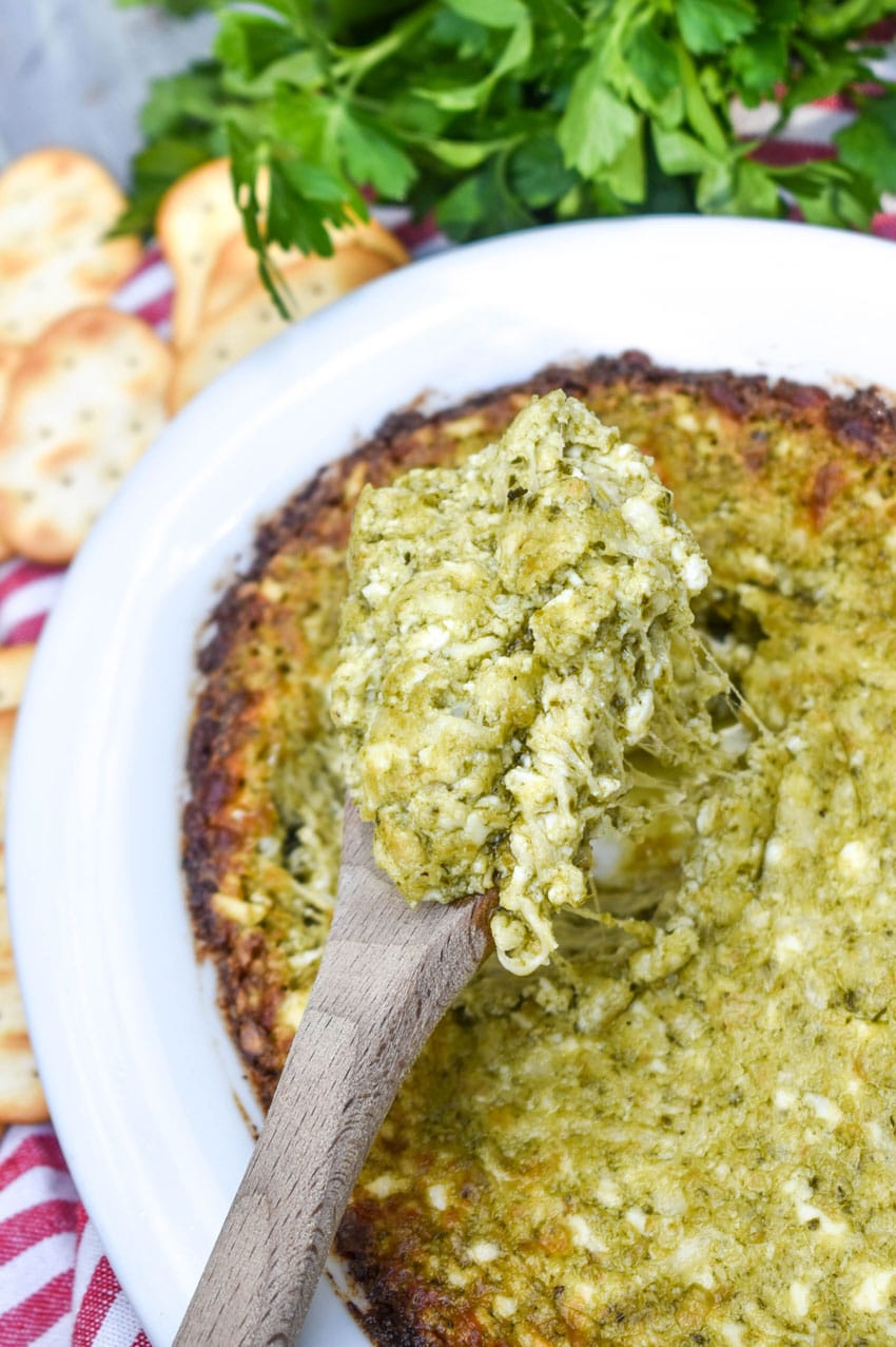 a wooden spoon scooping hot pesto dip out of a white serving dish