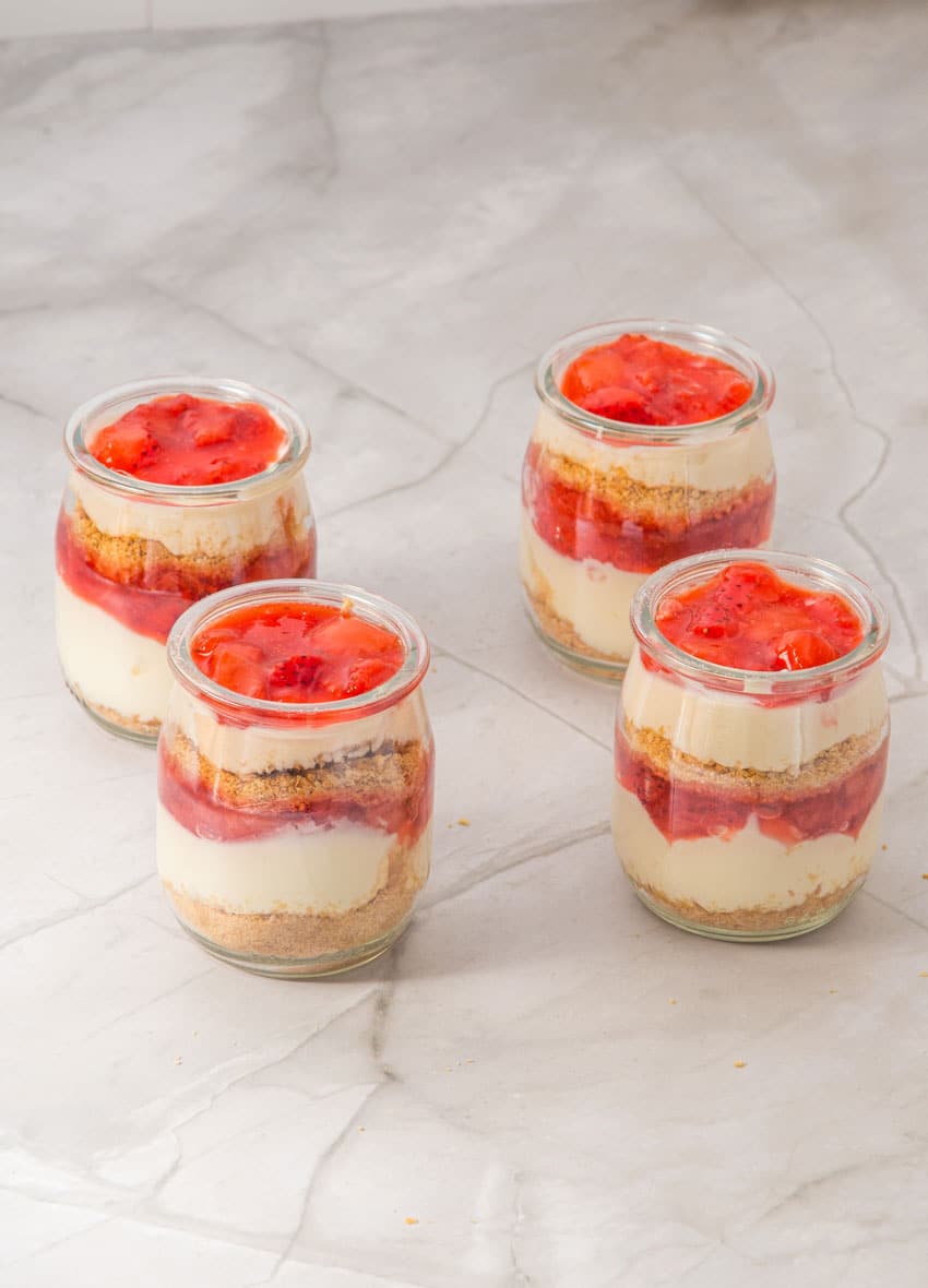 strawberry cheesecake layers being added to four small glass jars
