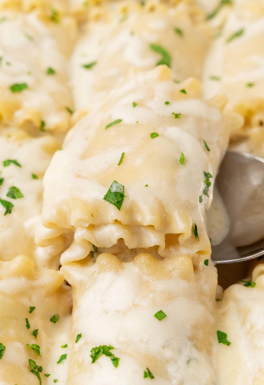 a silver spoon scooping a white chicken lasagna roll up out of a white baking dish