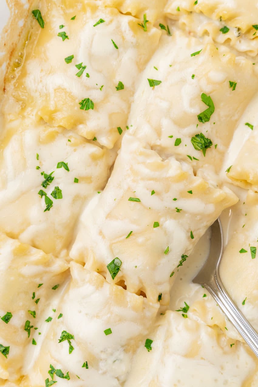 a silver spoon scooping a white chicken lasagna roll up out of a white baking dish