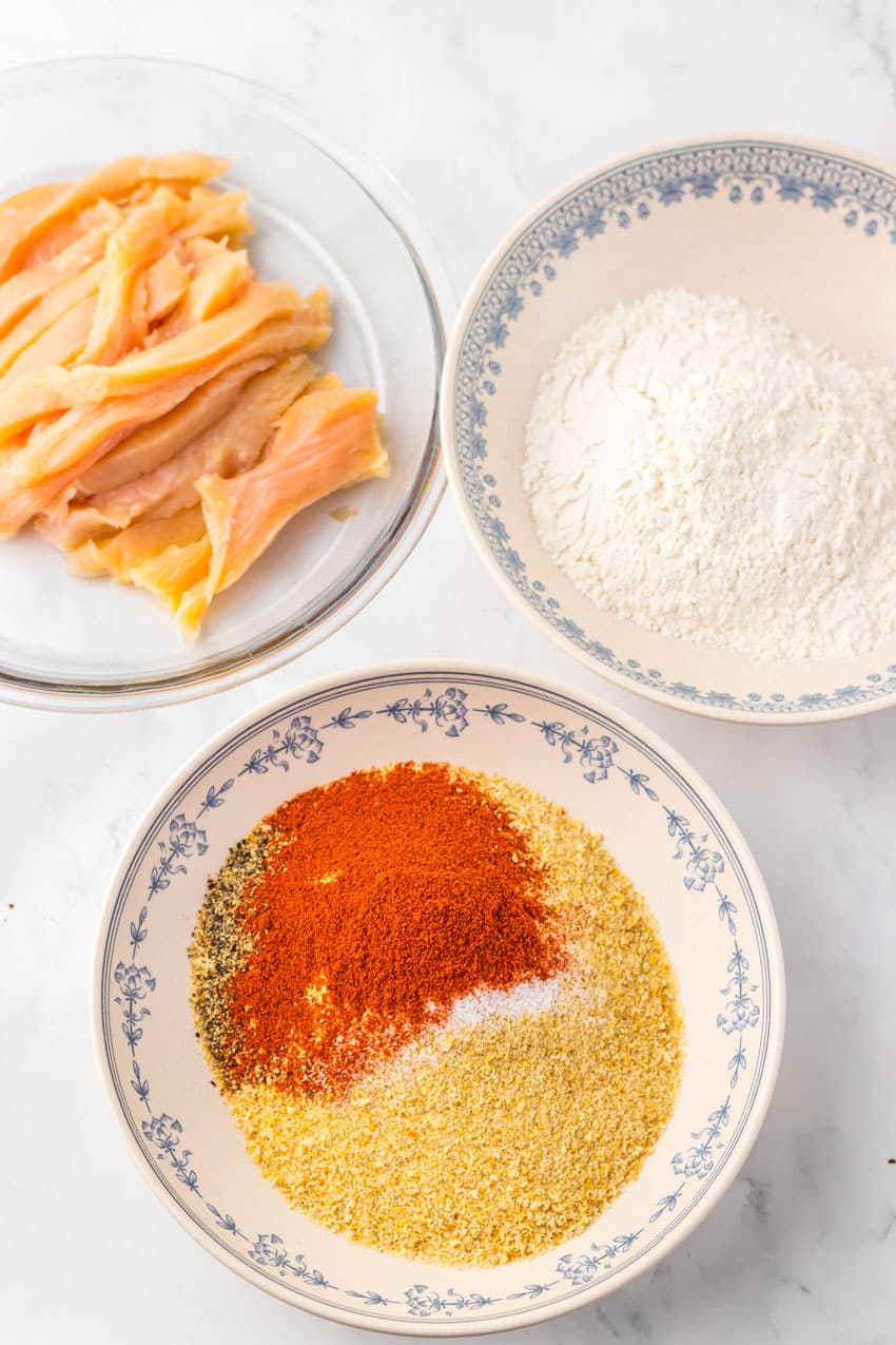 chicken strips on a gray plate ready to be dipped in a bowl of seasonings and another filled with flour