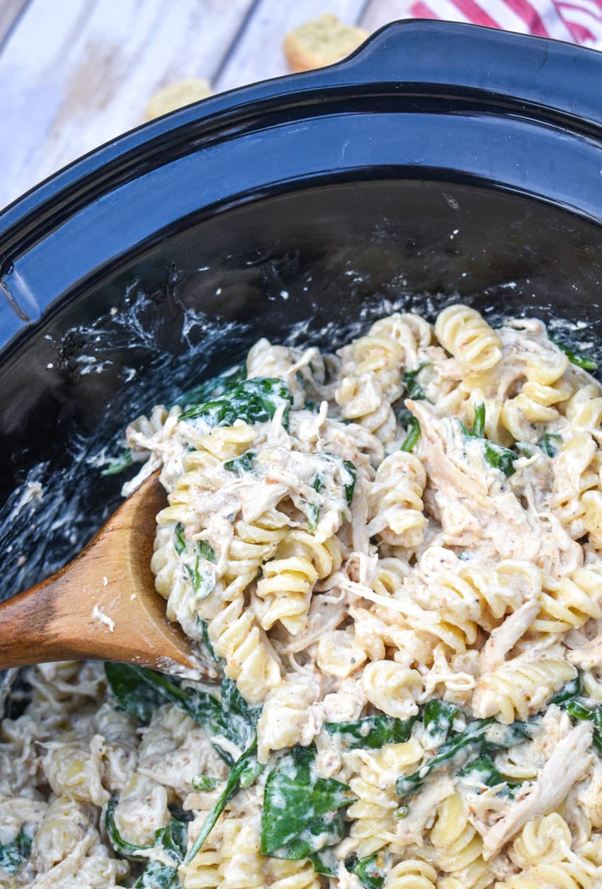 a wooden spoon scooping chicken caesar pasta out of the black bowl of a slow cooker