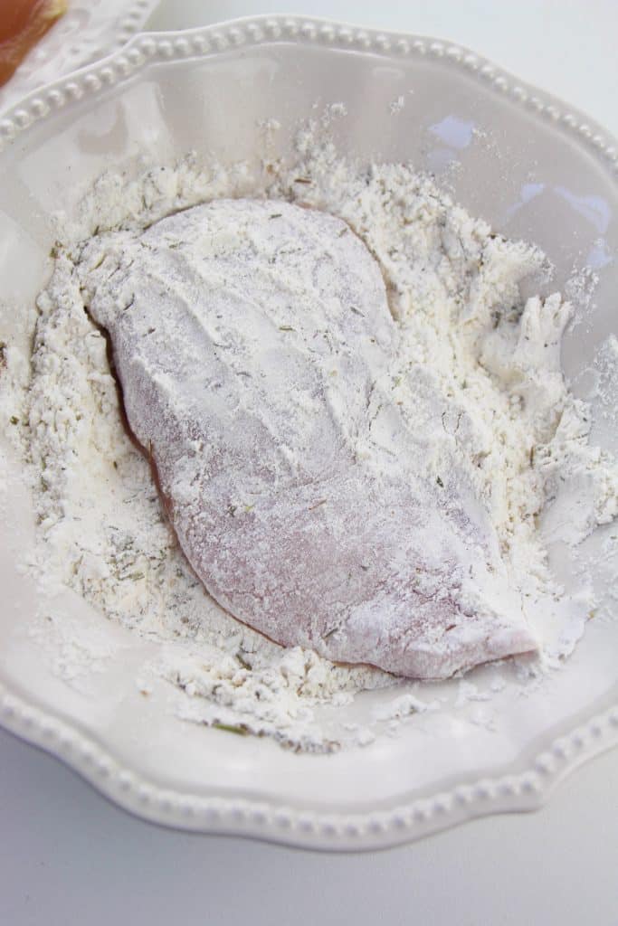 a chicken breast shown dredged in seasoned flour in a white bowl