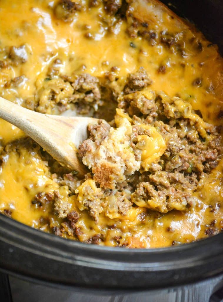 a wooden spoon scooping slow cooker cheeseburger casserole from a black crock