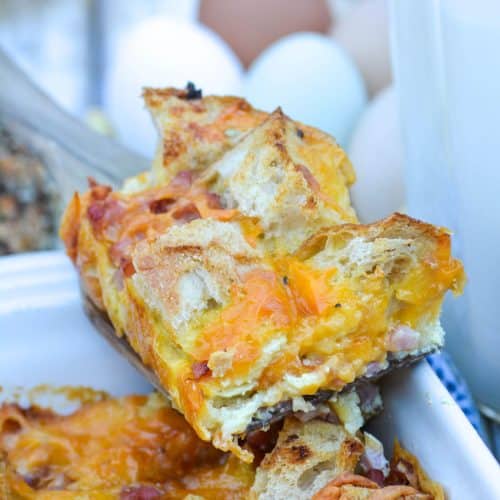 a slice of ham and cheese bread pudding on a wooden spatula resting on the edge of a casserole dish