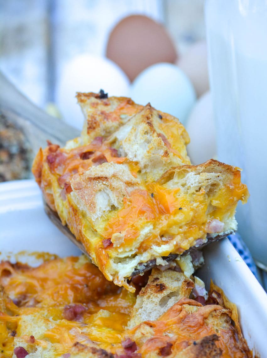 a slice of ham and cheese bread pudding on a wooden spatula resting on the edge of a casserole dish