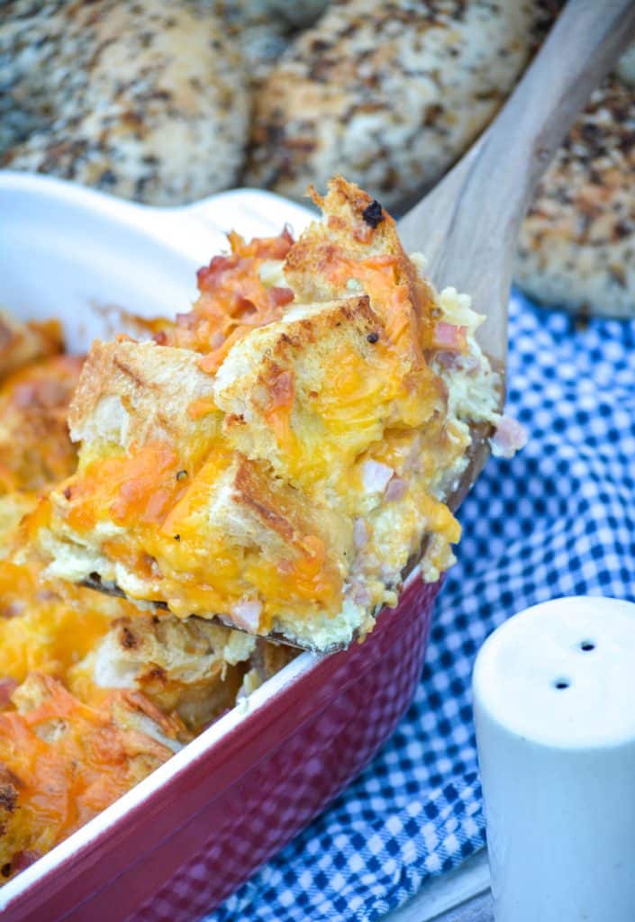 a slice of ham and cheese bread pudding on a wooden spatula resting on the edge of a casserole dish