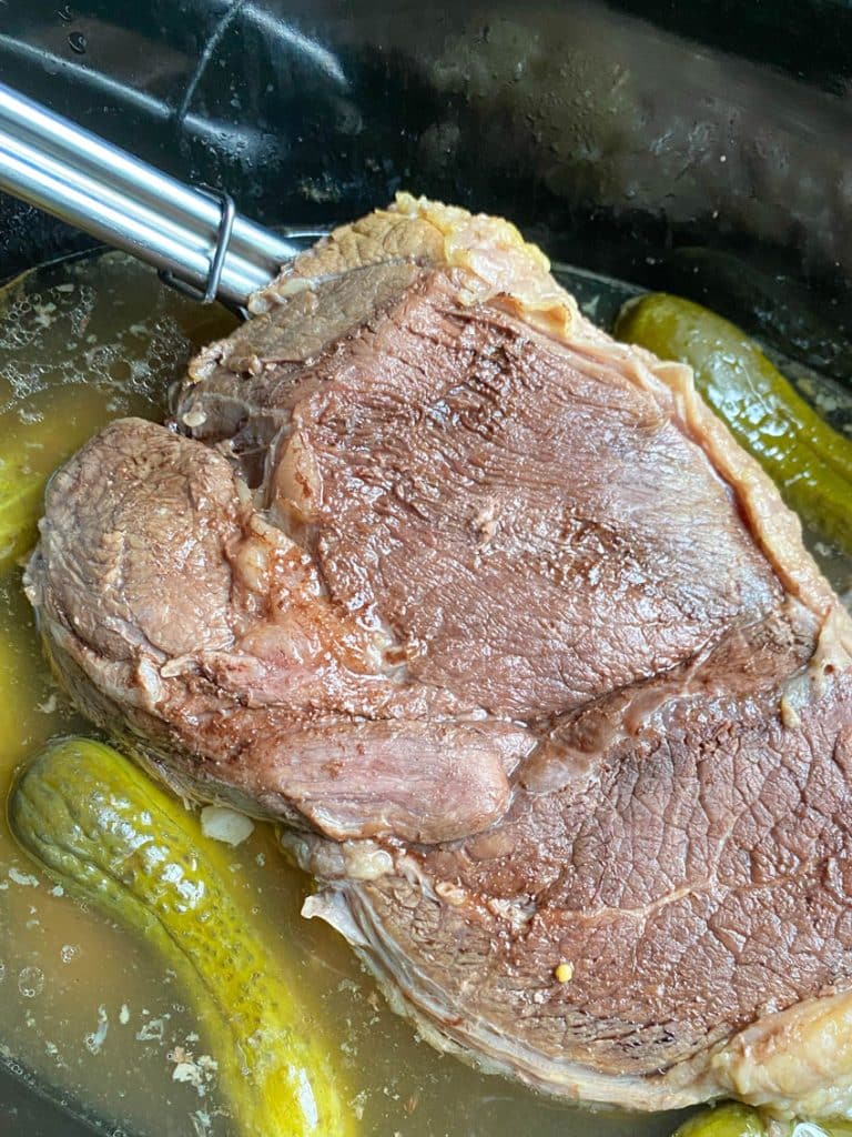 silver tongs holding up a cooked dill pickle pot roast in the black bowl of a crockpot