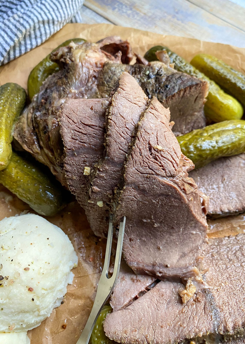 sliced slow cooker dill pickle pot roast with a meat fork on a parchment paper lined cutting board