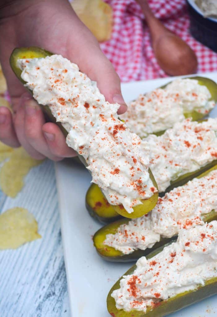 a hand holding a chicken salad stuffed pickle over a platter of prepared pickles