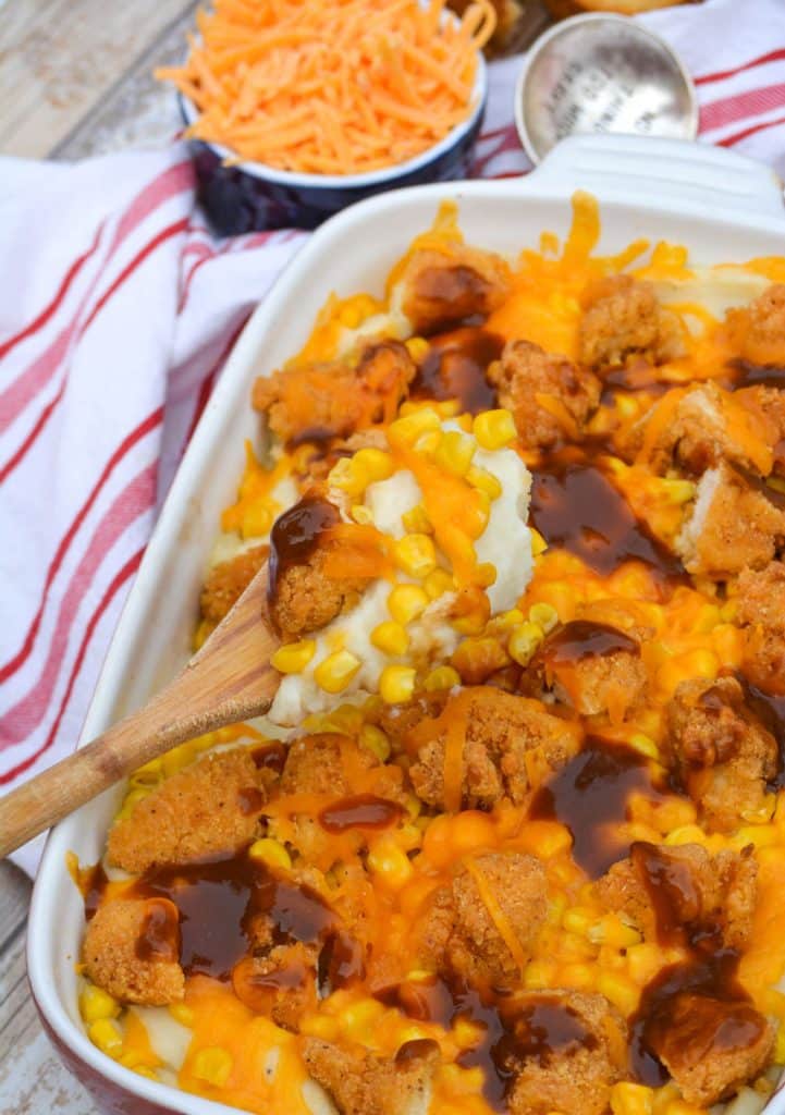 a wooden spoon holding up a scoop of mashed potatoes topped with shredded cheese, corn kernels, crisp chicken, and brown gravy