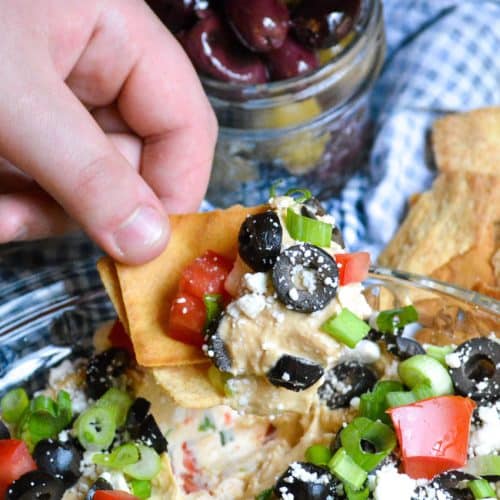a hand holding up a pita chip topped with greek dip