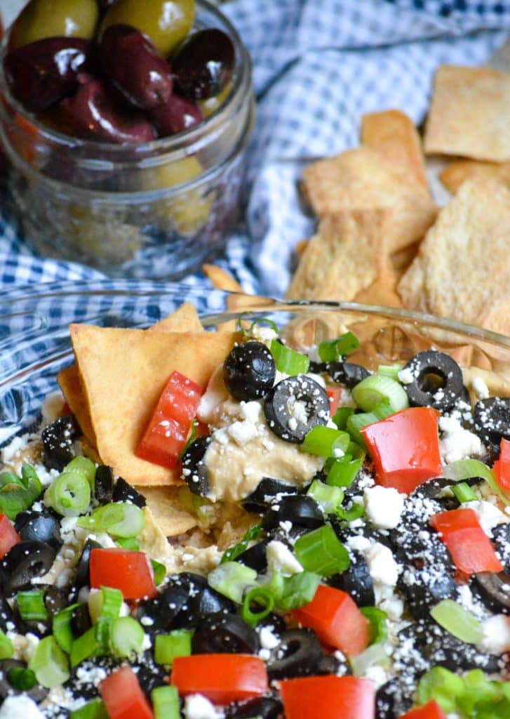 a square pita cracker dipped in a dish of layered greek dip