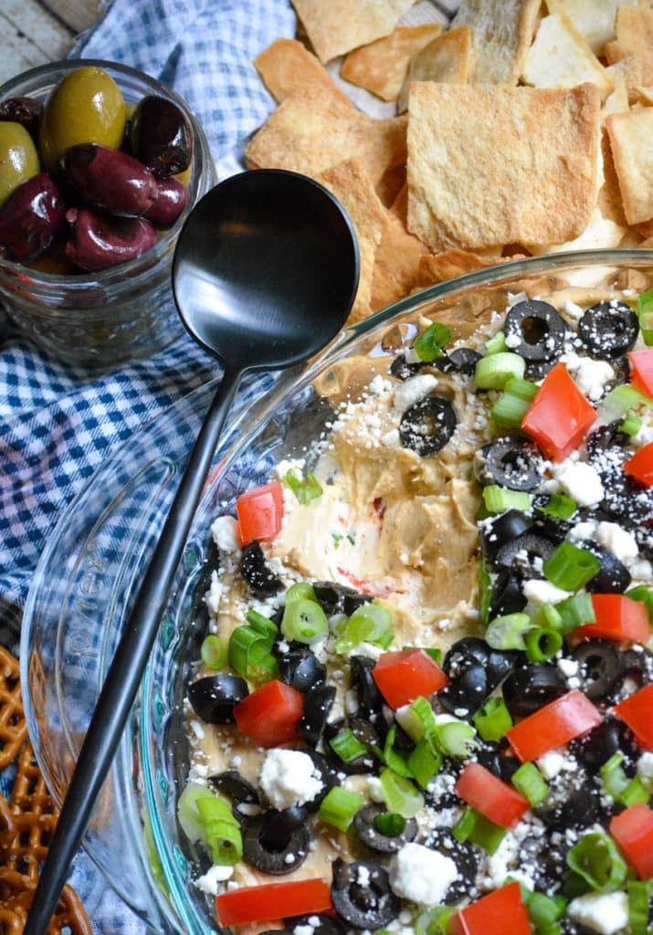 a black spoon resting on a glass dish filled with greek layer dip