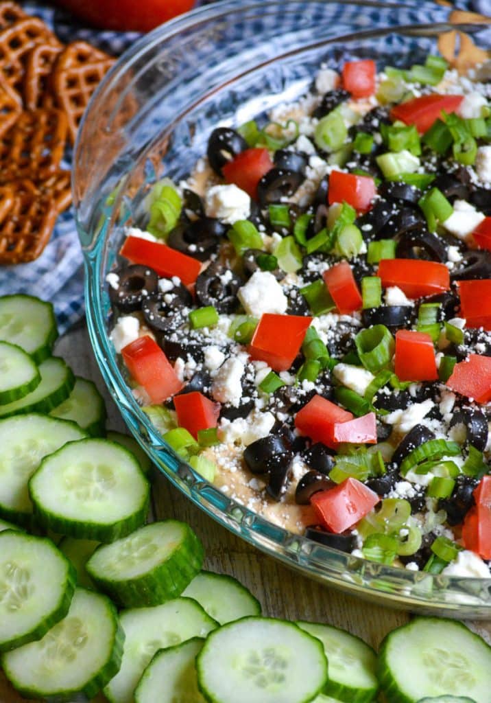 7 layer Greek dip in a glass pie pan surrounded by crackers and pretzels