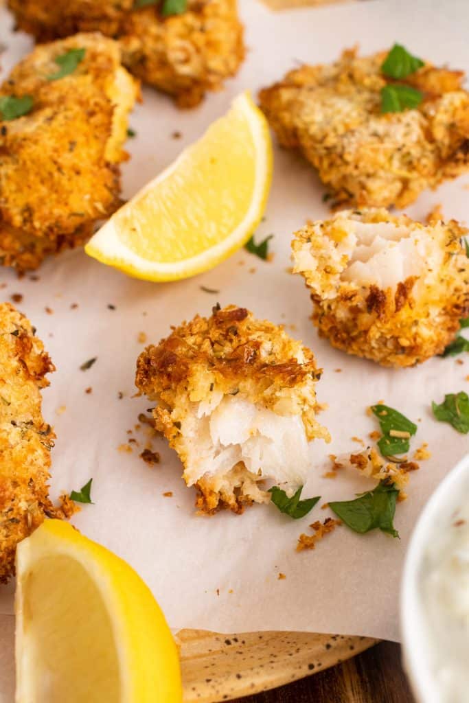 air fryer fish sticks on a parchment paper covered cutting board with fresh herbs and lemon wedges