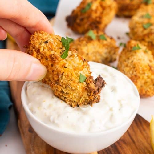 a hand dipping an air fryer fish stick into a white bowl of tartar sauce