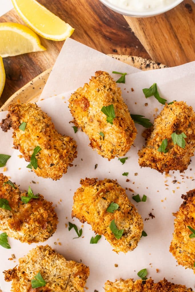 air fryer fish sticks on a parchment paper covered cutting board with fresh herbs and lemon wedges