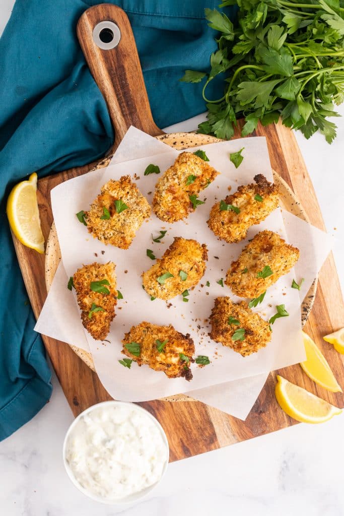 air fryer fish sticks on a parchment paper covered cutting board with fresh herbs and lemon wedges