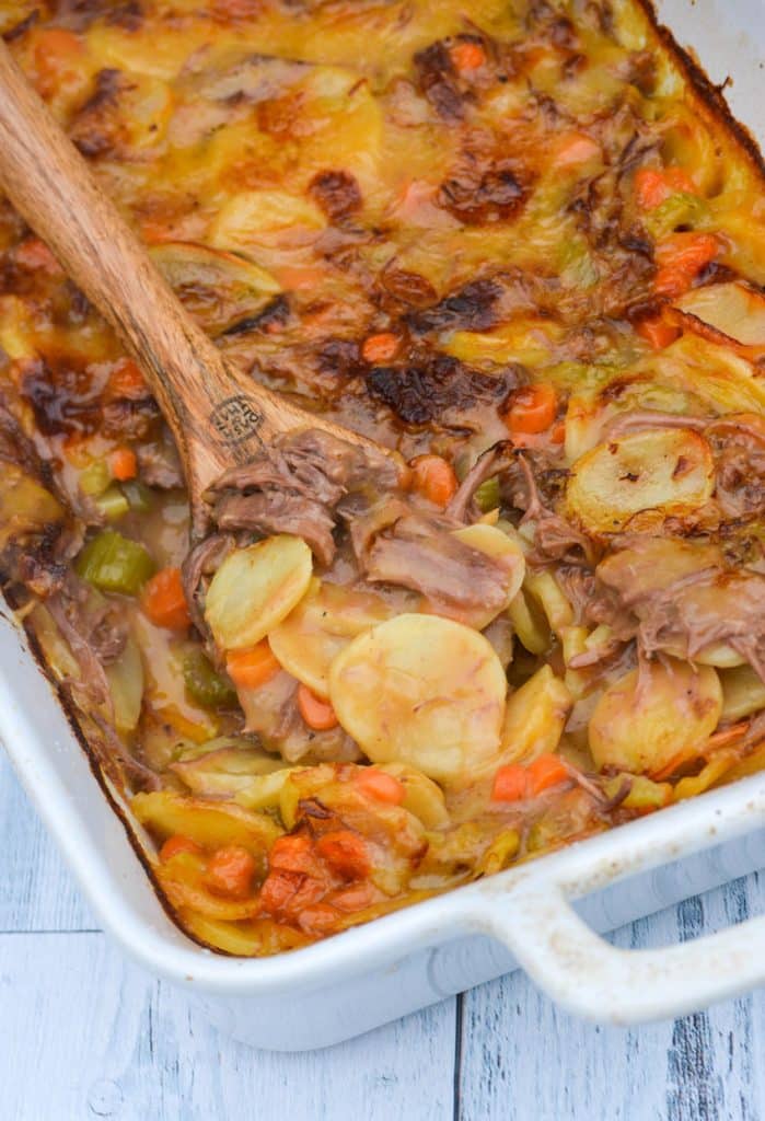 a wooden spoon digging into a white casserole dish filled with cheesy potatoes & leftover pot roast