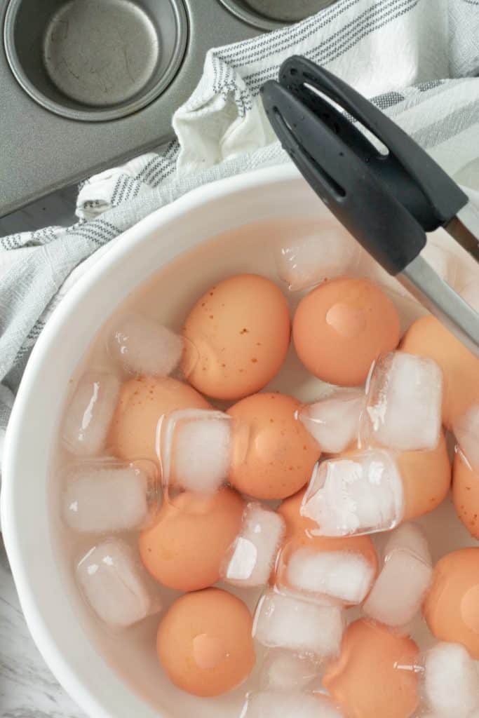 oven baked hard boiled eggs in an ice bath with a pair of tongs resting on the edge of the bowl