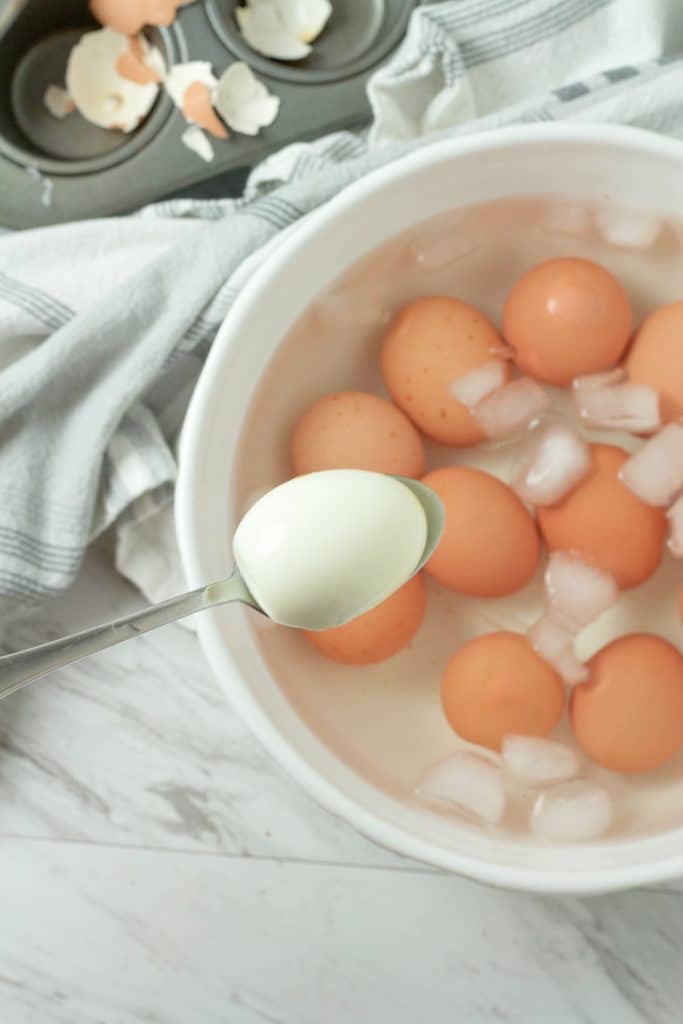 a silver spoon holding a peeled baked hard boiled egg over eggs in an ice bath