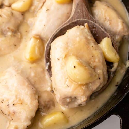 a wooden spoon resting in a black skillet holding a chicken thigh in garlic gravy