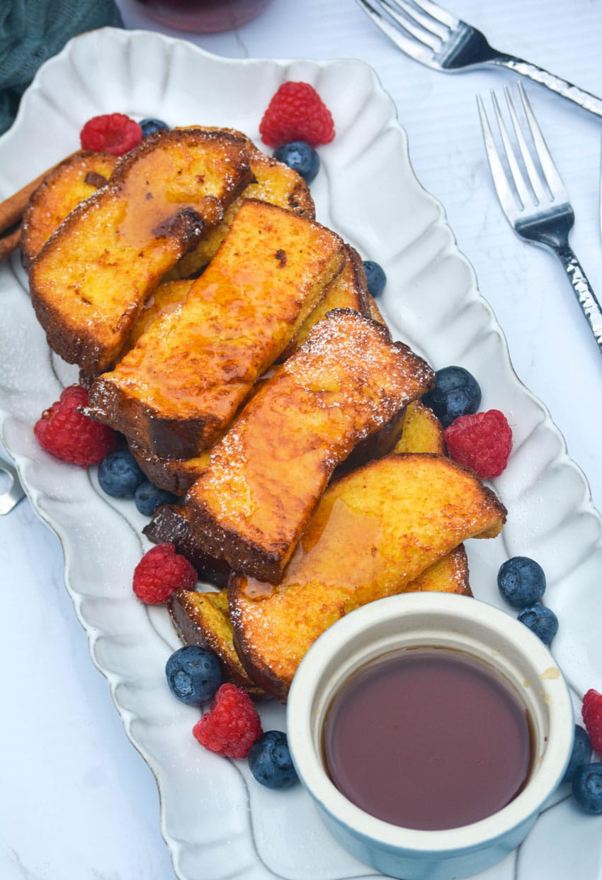 air fryer french toast sticks on a white platter surrounded by fresh berries with a small bowl of syrup on the side