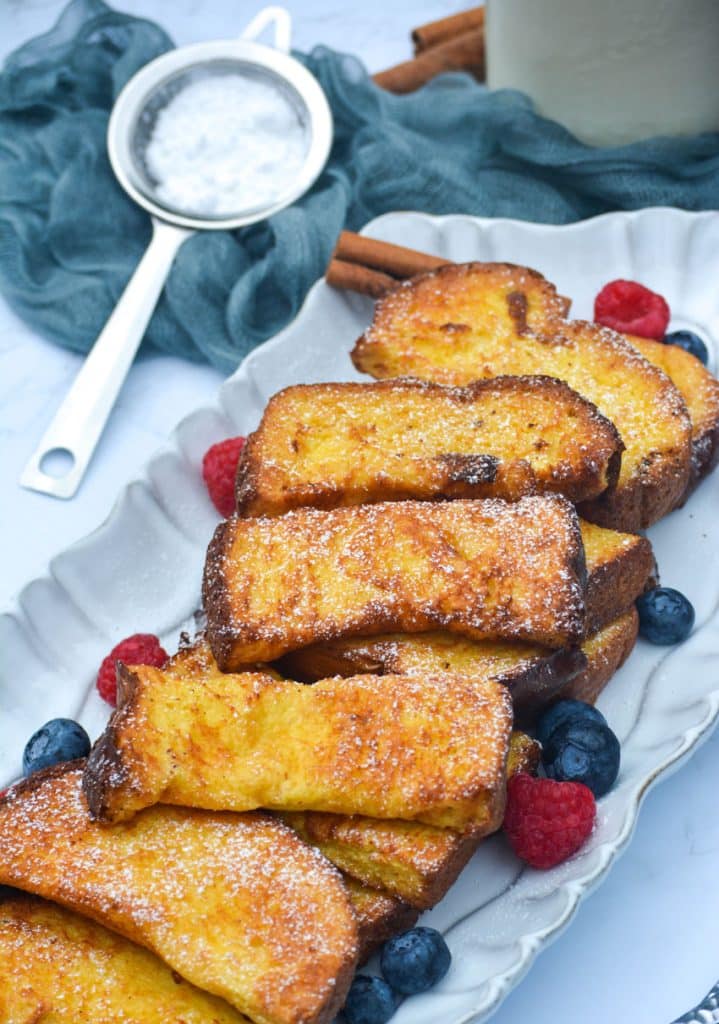 air fryer french toast sticks on a white platter surrounded by fresh berries with a sifter of powdered sugar on the side
