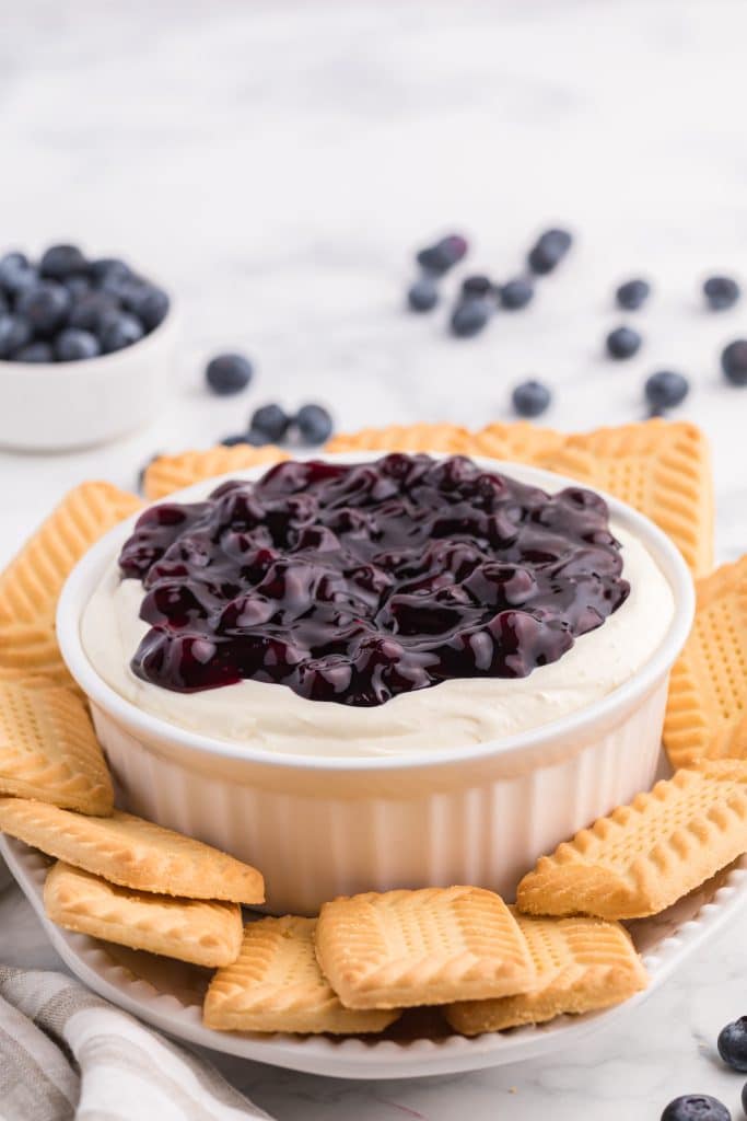 a white bowl filled with blueberry cheesecake dip surrounded by shortbread cookies