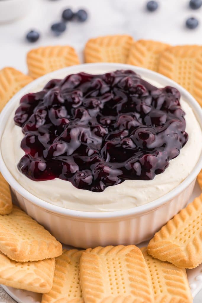a white bowl filled with blueberry cheesecake dip surrounded by shortbread cookies