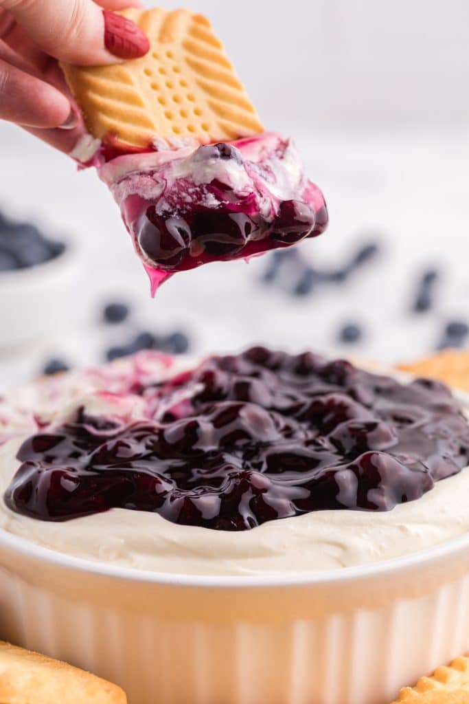 a hand holding up a shortbread cookie with blueberry cheesecake dip on it