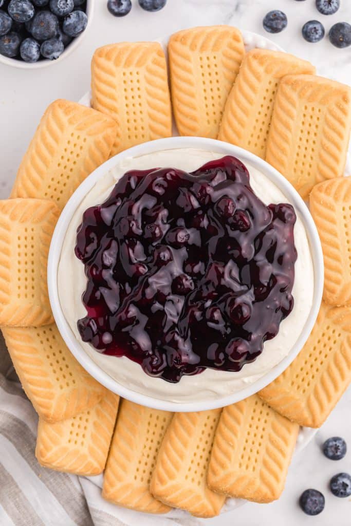 a white bowl filled with blueberry cheesecake dip surrounded by shortbread cookies