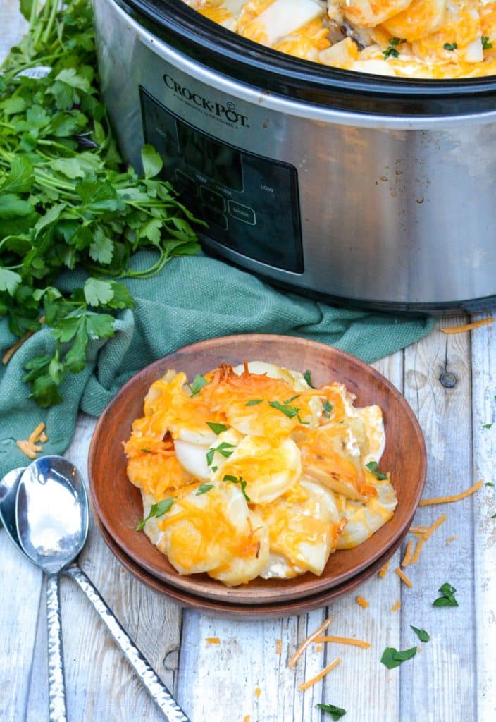 cheesy crockpot scalloped potatoes on a wooden plate in front of a silver slow cooker