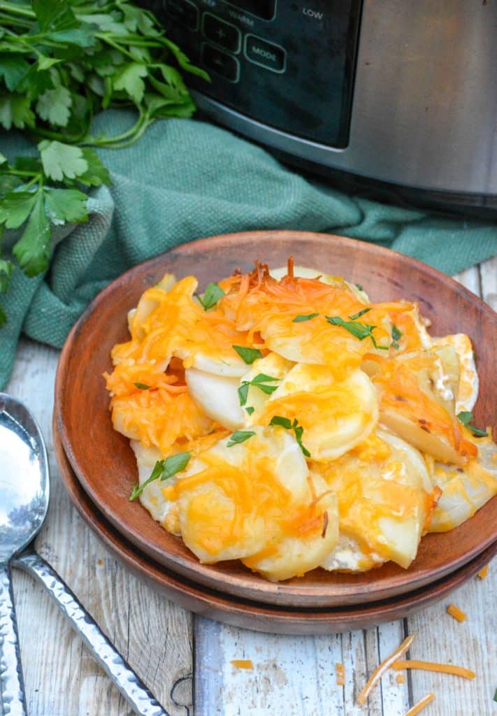 cheesy crockpot scalloped potatoes on a wooden plate in front of a silver slow cooker