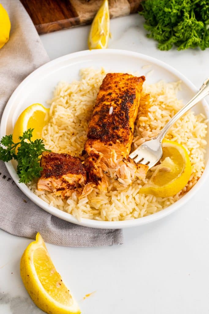 a fork holding a piece of blackened salmon on a bed of white rice on a white plate