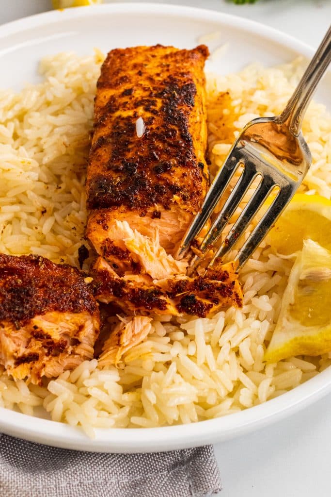 a silver fork flaking apart a blackened salmon filet resting on a bed of white rice