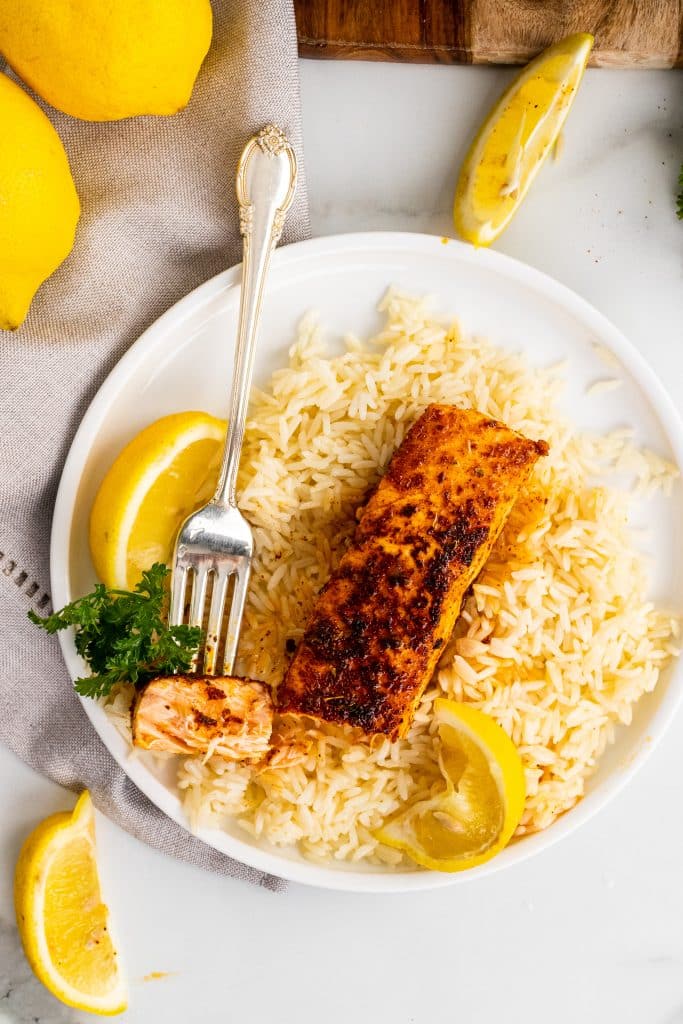 a fork holding a piece of blackened salmon on a bed of white rice on a white plate