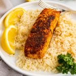 a blackened salmon filet on a bed of white rice next to lemon wedges on a white plate